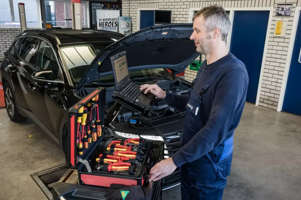Monteur leest met een laptop een auto uit, met de gereedschapskoffer open naast de motorkap